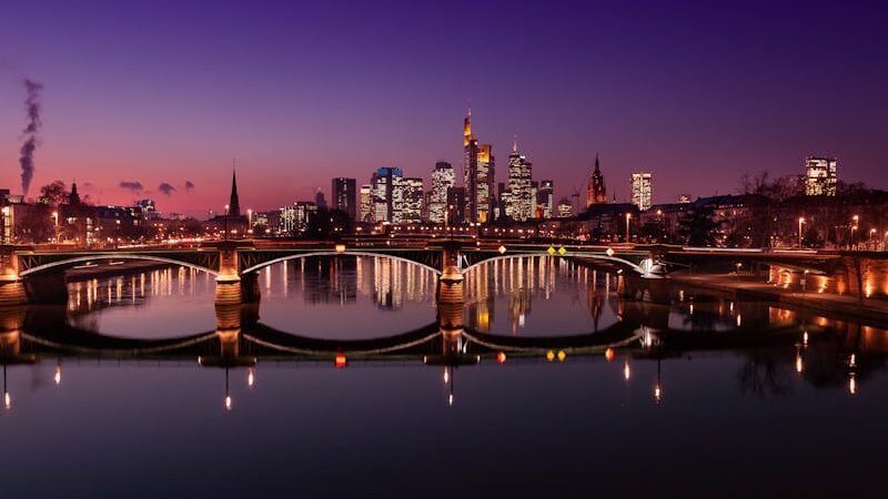 **"Night view of Frankfurt, Germany, with a brightly lit bridge reflecting on the Main River and city skyscrapers in the background."**
